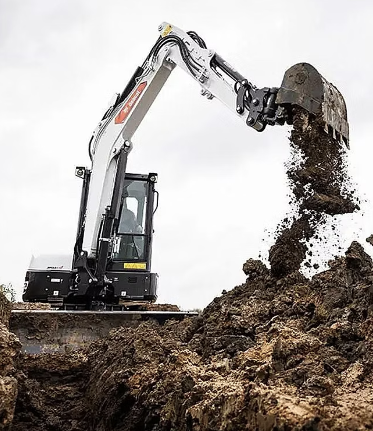 A small excavator digging dirt in a field, with soil being displaced and a clear blue sky in the background.