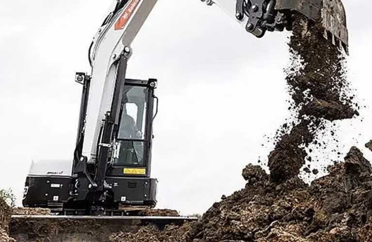 A small excavator digging dirt in a field, with soil being displaced and a clear blue sky in the background.