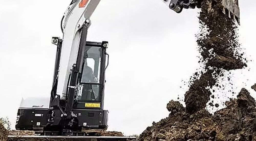A small excavator digging dirt in a field, with soil being displaced and a clear blue sky in the background.